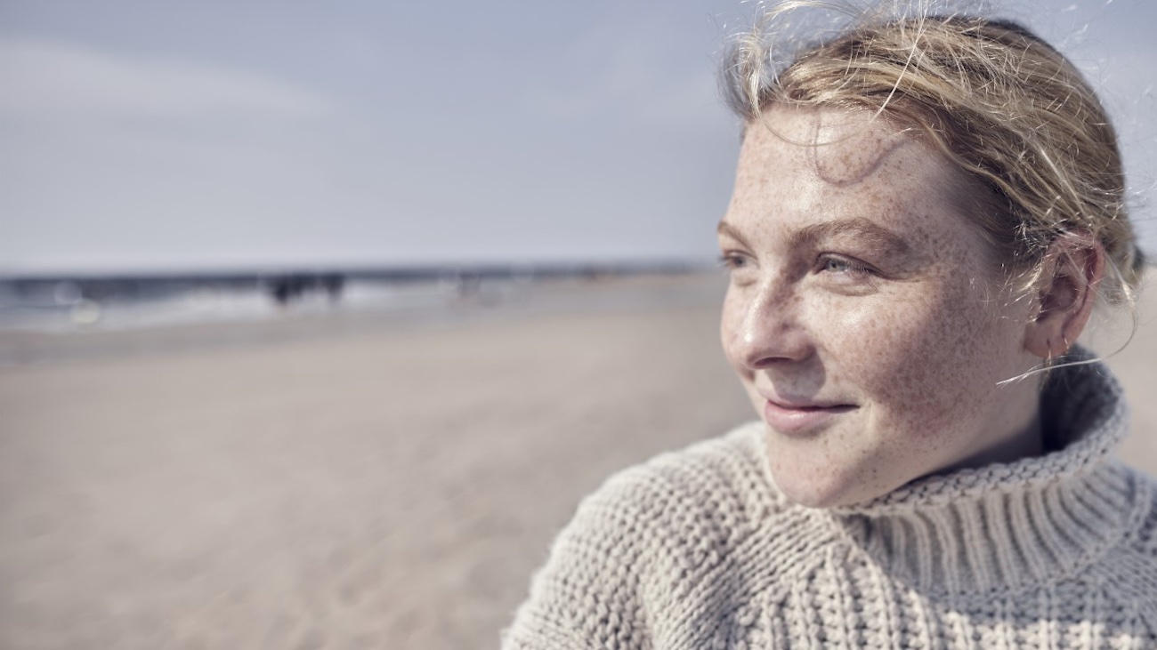 En ung kvinne med fregner og kort, bølget hår smiler mens hun ser ut mot havet. Hun har på seg en lys, tykk genser og står på en strand, med en klar himmel og mennesker i bakgrunnen.